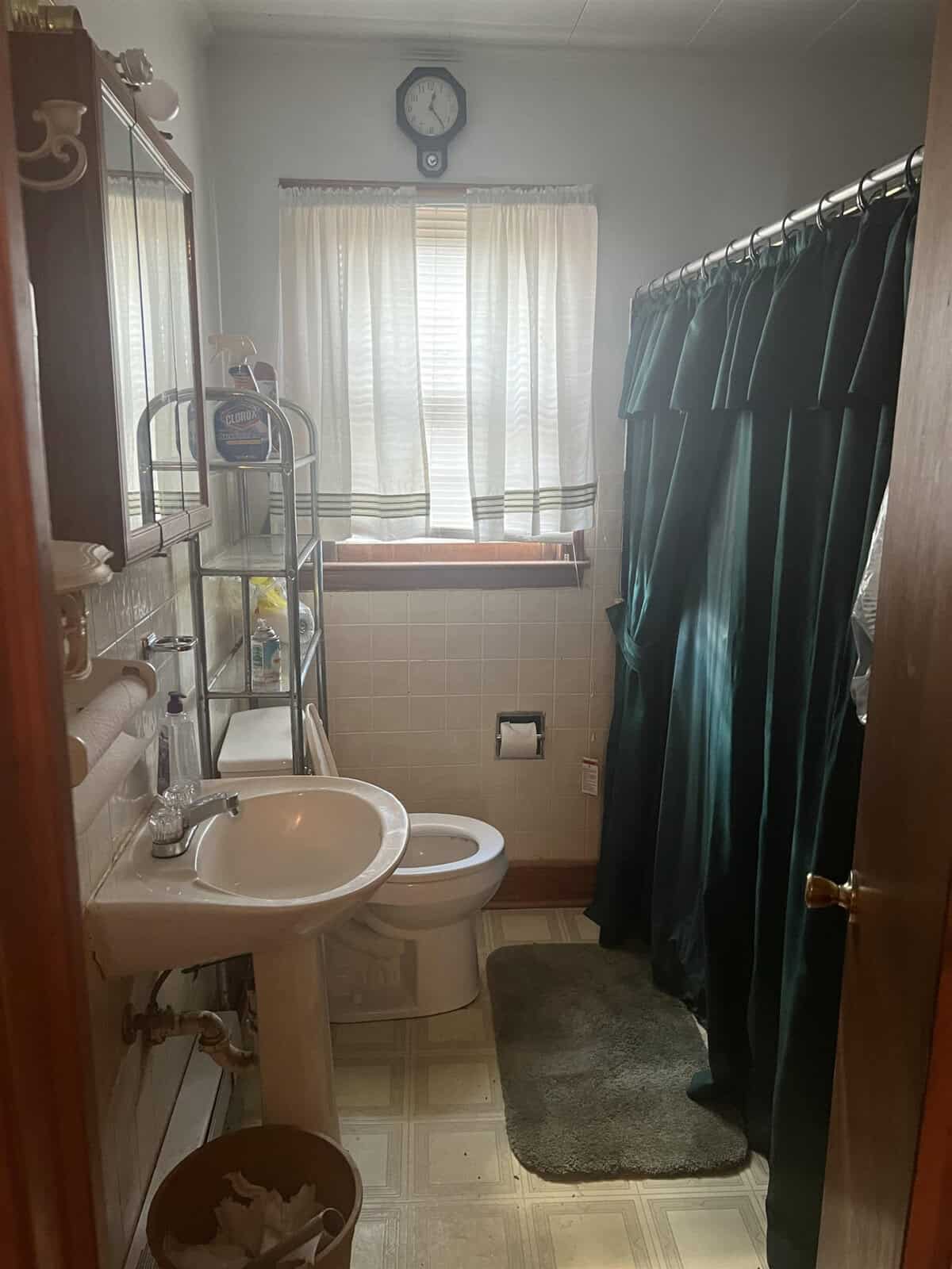 A bathroom with a toilet, sink, and shower curtain, featuring a window with curtains, a wall clock, and various bath accessories.