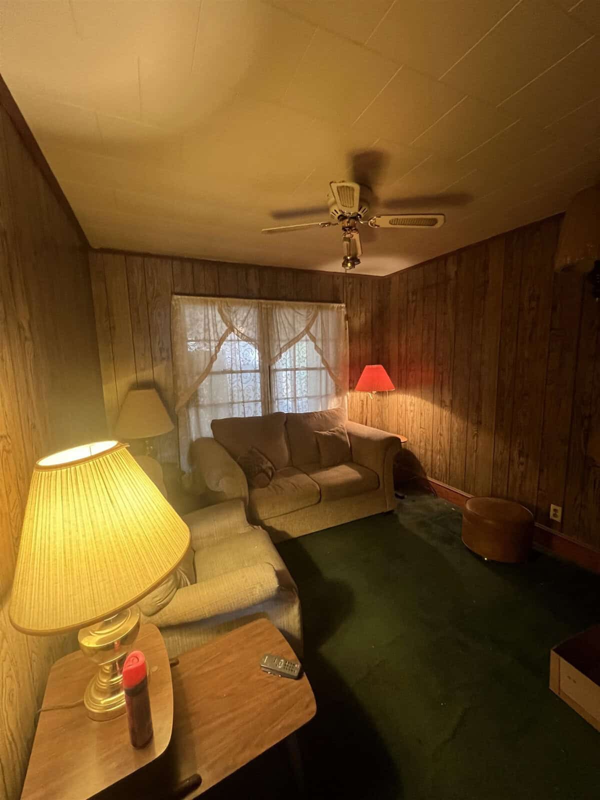 A cozy, dimly lit room featuring wood paneling, a sofa with throw pillows, two lamps with illuminated shades, and a ceiling fan.