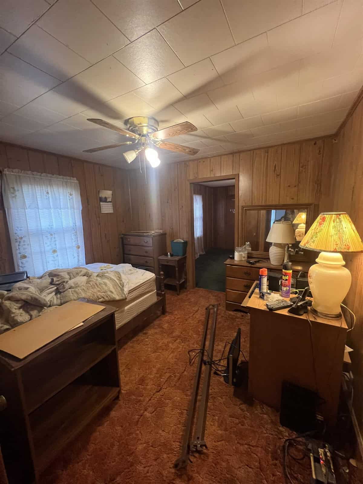 A vintage-style bedroom featuring wood-paneled walls, a shaggy orange carpet, and a ceiling fan. the room contains a bed with an unmade mattress, a wooden dresser, a small desk, a nightstand with a lamp, and various personal items scattered throughout.