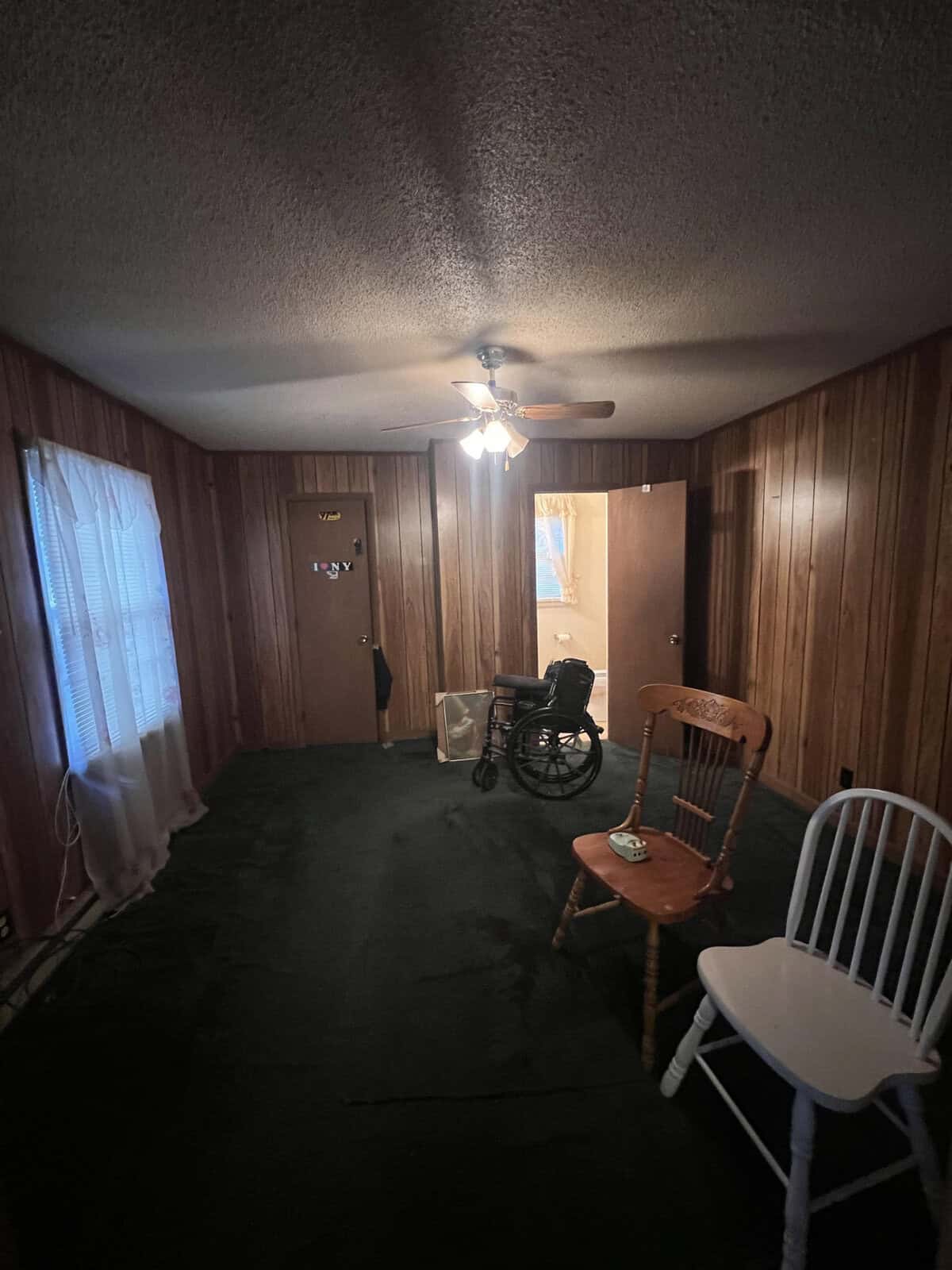 An interior room featuring wood-paneled walls, two chairs, a ceiling fan with a light, a black floor, and a wheelchair positioned near the center of the room. the room has a window dressed with sheer curtains and a door partially open.