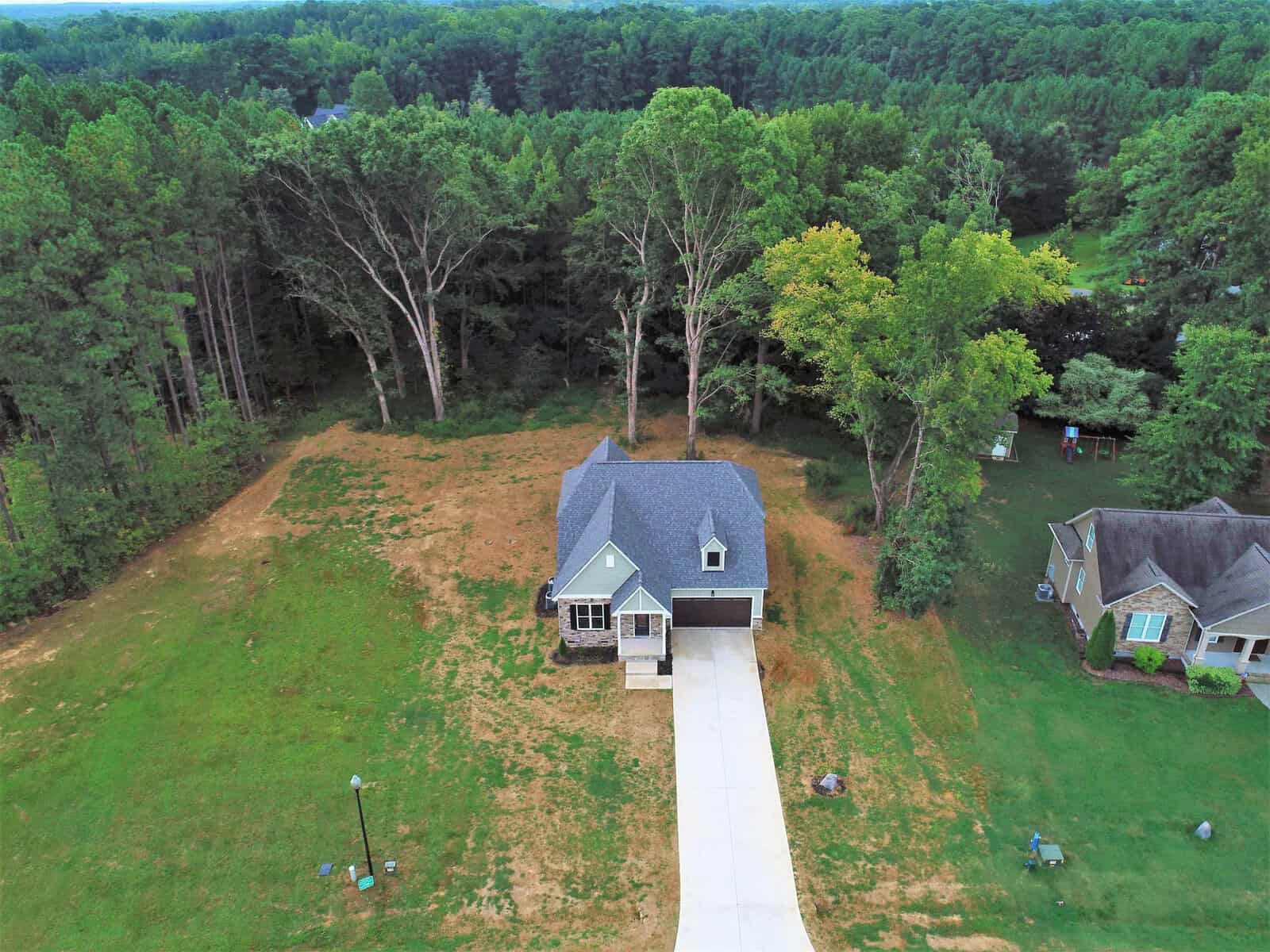 Aerial view of a gray house with a steep roof, a driveway, and a garage. It's surrounded by a large lawn and tall trees, with a dense forest in the background. Nearby, another house is partially visible to the right.
