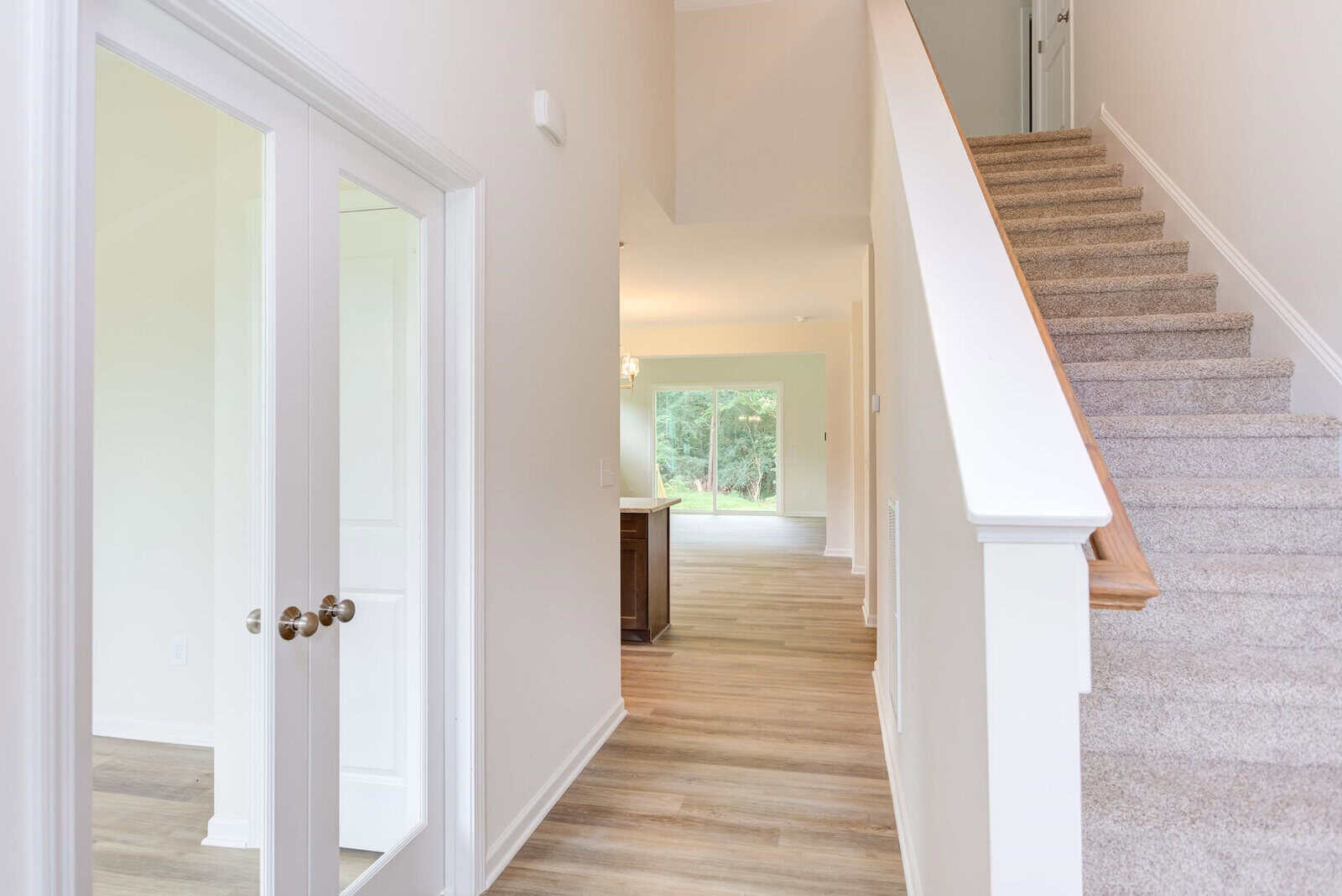 A bright and spacious entryway features a staircase with carpeted steps on the right, leading upstairs. On the left, there's a set of double glass doors. The corridor extends towards a living area with large windows overlooking greenery.