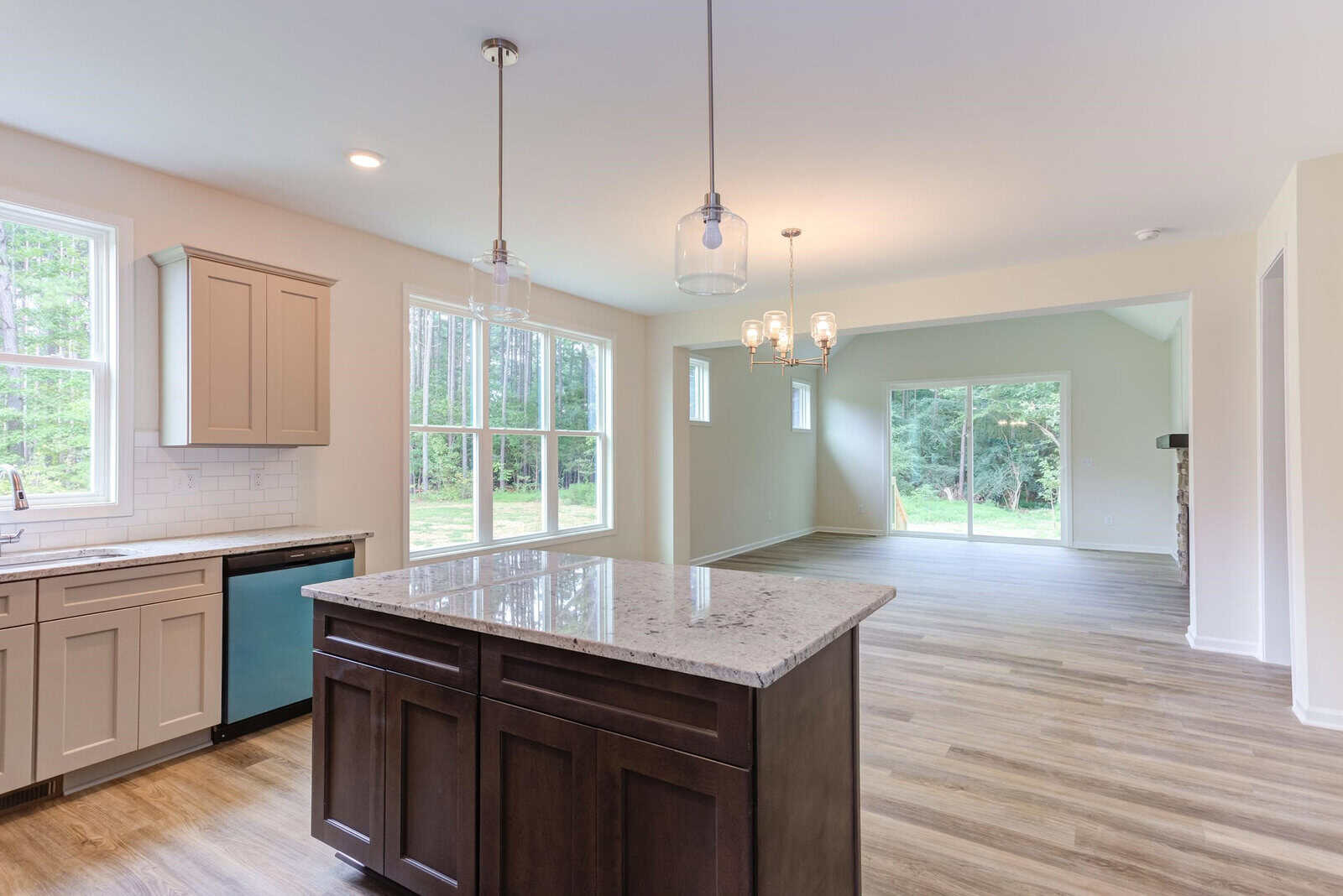 Bright open-plan kitchen with large windows. Features an island with a granite countertop, beige cabinets, a dishwasher, and light wood flooring. Adjacent dining area with chandelier and sliding glass doors leading outside.