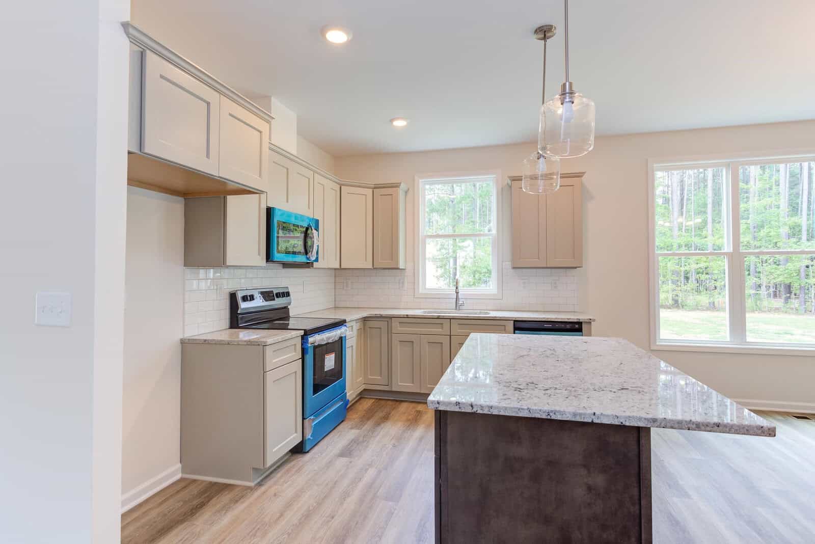 A modern kitchen with light gray cabinets, stainless steel appliances, and a large island with a granite countertop. The room features wooden flooring and two pendant lights, with natural light streaming in through the large windows.