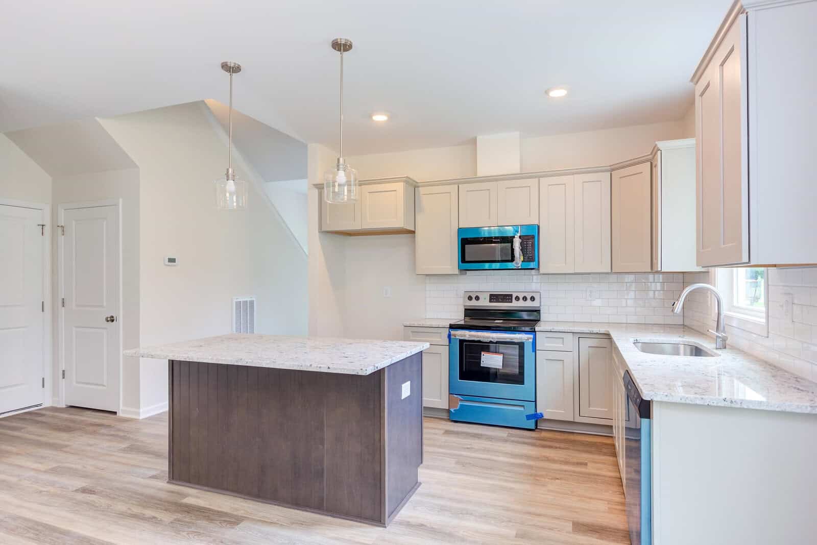 A modern kitchen with white cabinets, a granite countertop island, and stainless steel appliances, including an oven and microwave. Pendant lights hang above the island, and the floor is light wood.