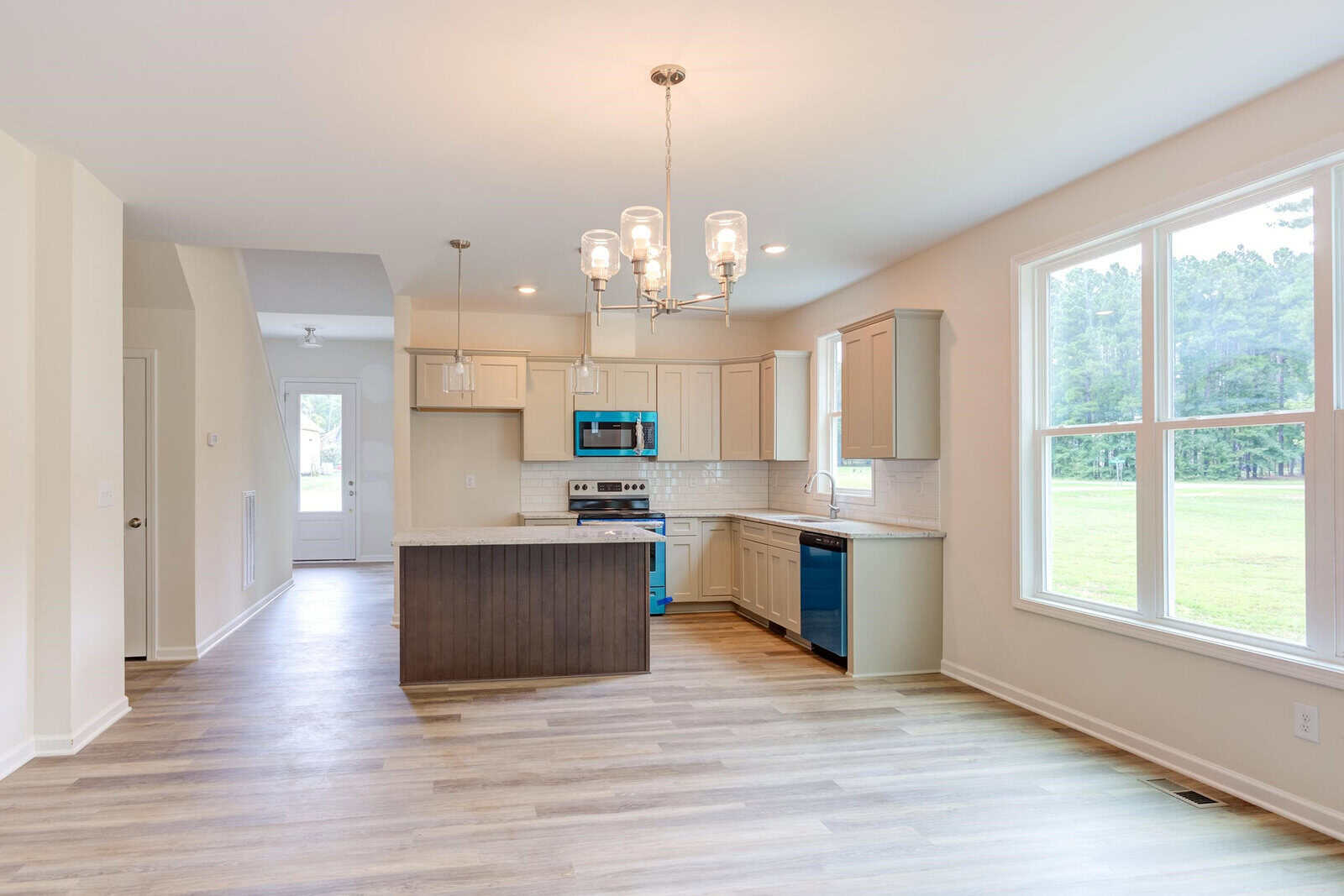 A modern kitchen with light wood flooring, a central island, white cabinets, stainless steel appliances, and a large window. Overhead, a stylish chandelier provides lighting. The room is bright and airy, with a door visible in the background.