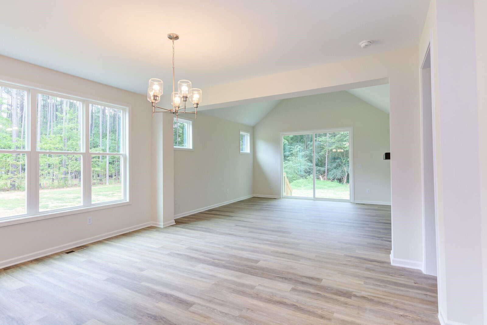Bright, empty room with light wooden flooring, large windows, and a glass sliding door leading to an outdoor area. A modern chandelier hangs from the ceiling, casting soft light across the space. Walls are painted light cream.