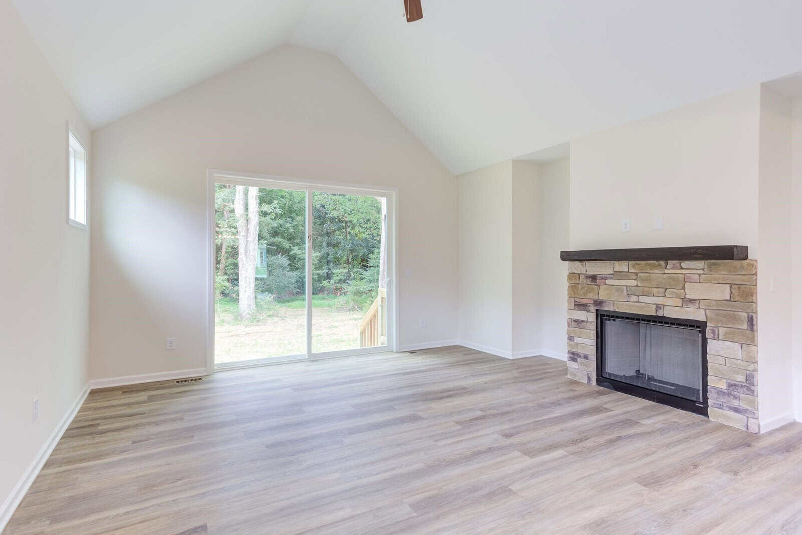 A bright, empty living room with vaulted ceilings features a sliding glass door opening to a wooded view. The room has light wood flooring and a stone fireplace with a dark wooden mantel on the right. Walls are painted white.