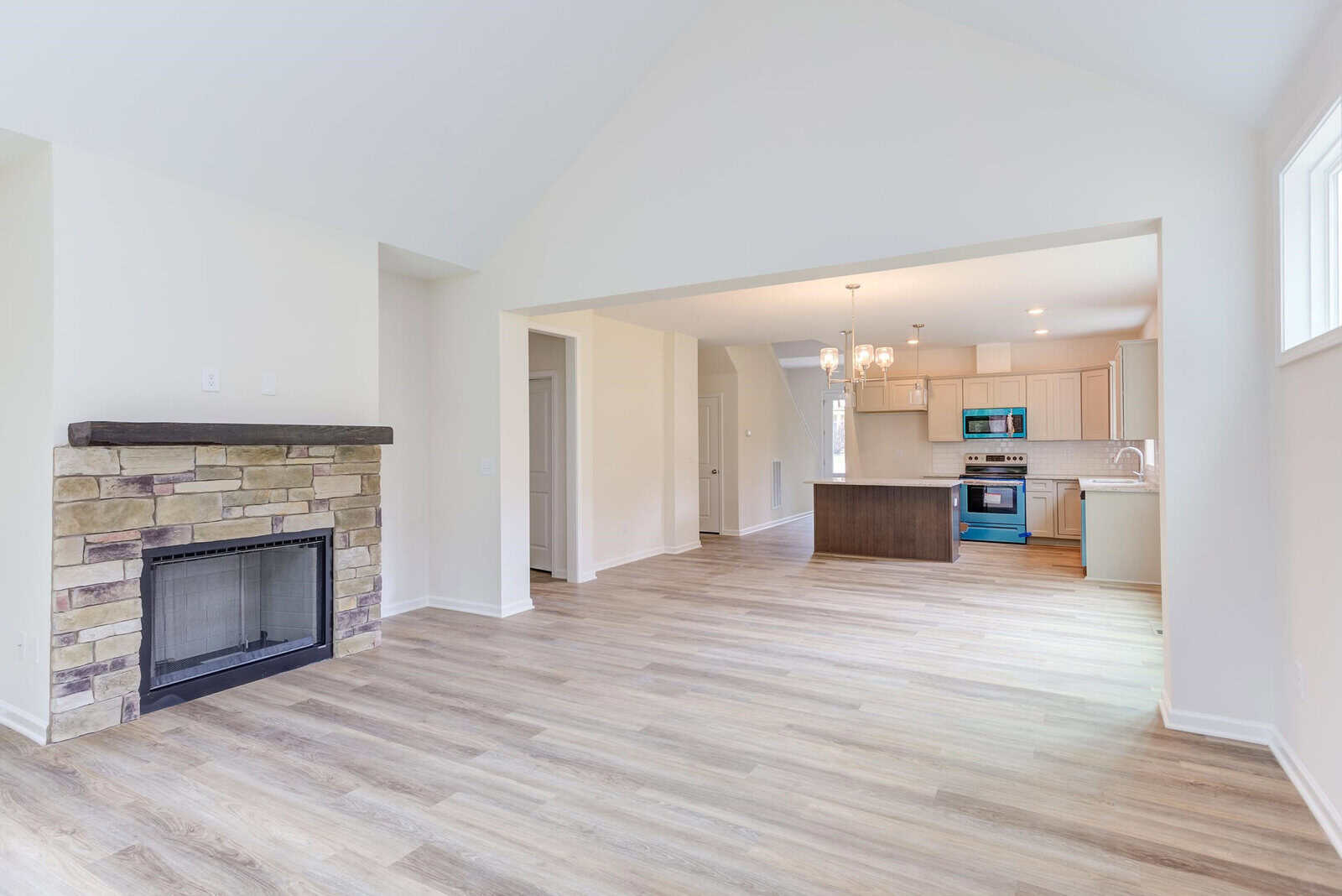 Spacious open-plan interior with light wood flooring and neutral walls. Features a stone fireplace, modern kitchen with light cabinets, blue appliances, and a central island. Large windows provide natural light.