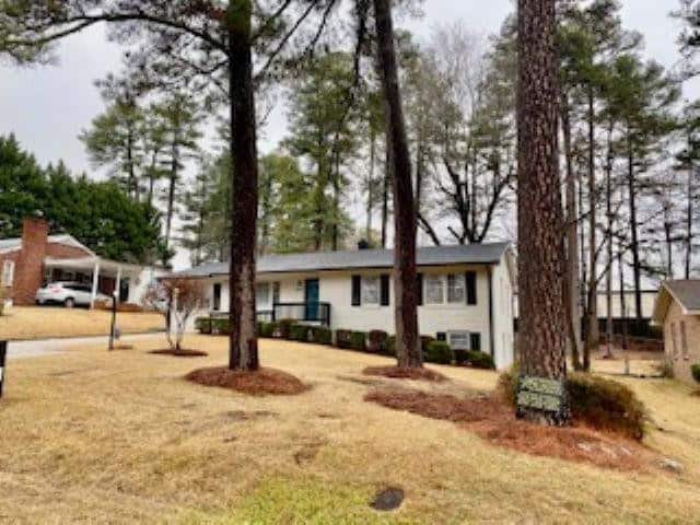 A single-story white house with a dark roof, surrounded by tall pine trees, situated on a sloping, grassy lawn with a partly cloudy sky overhead.