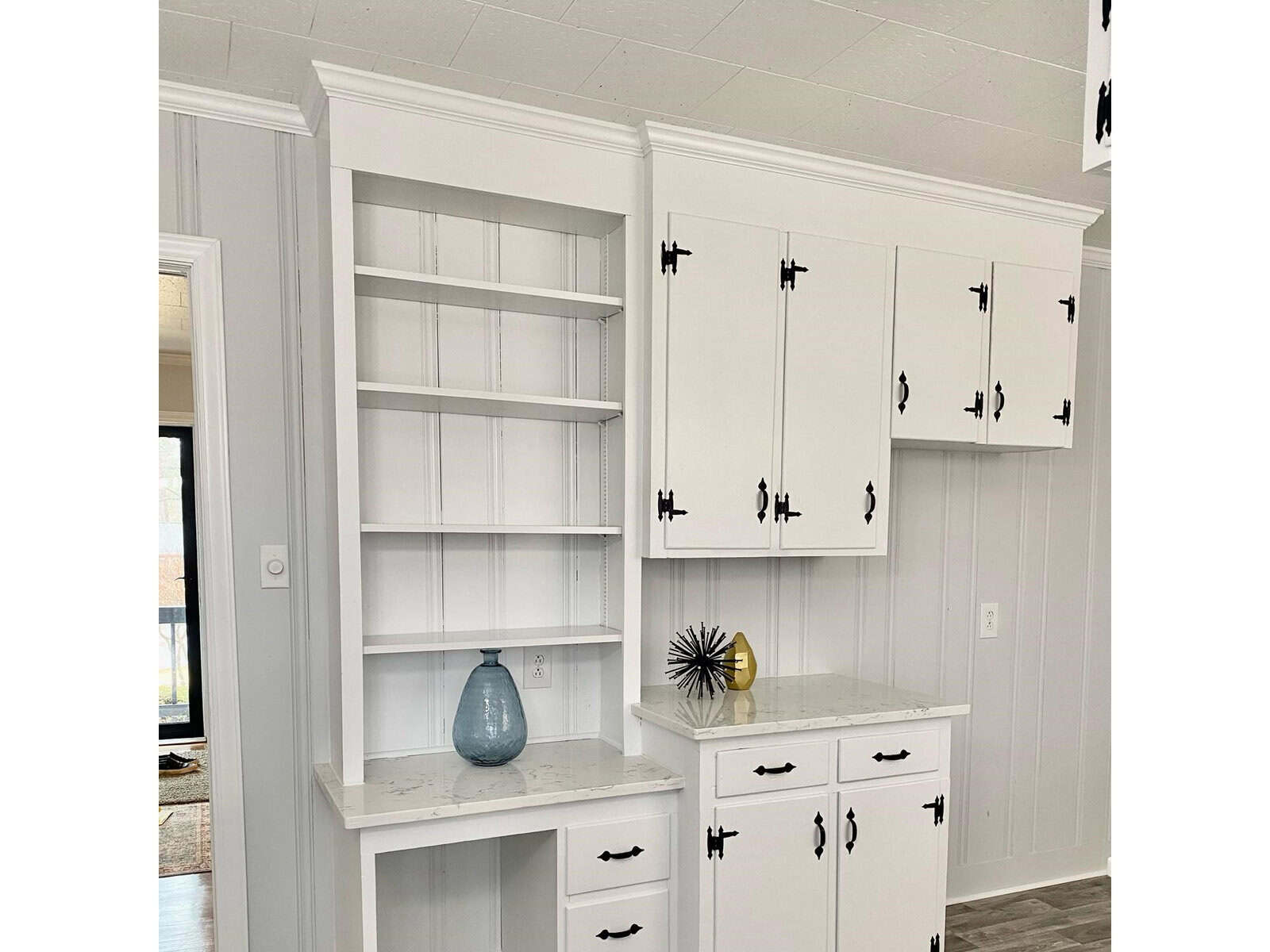 A white, modern kitchen interior with empty shelves, cabinets with black handles, and a countertop displaying a blue vase and a small decorative plant.