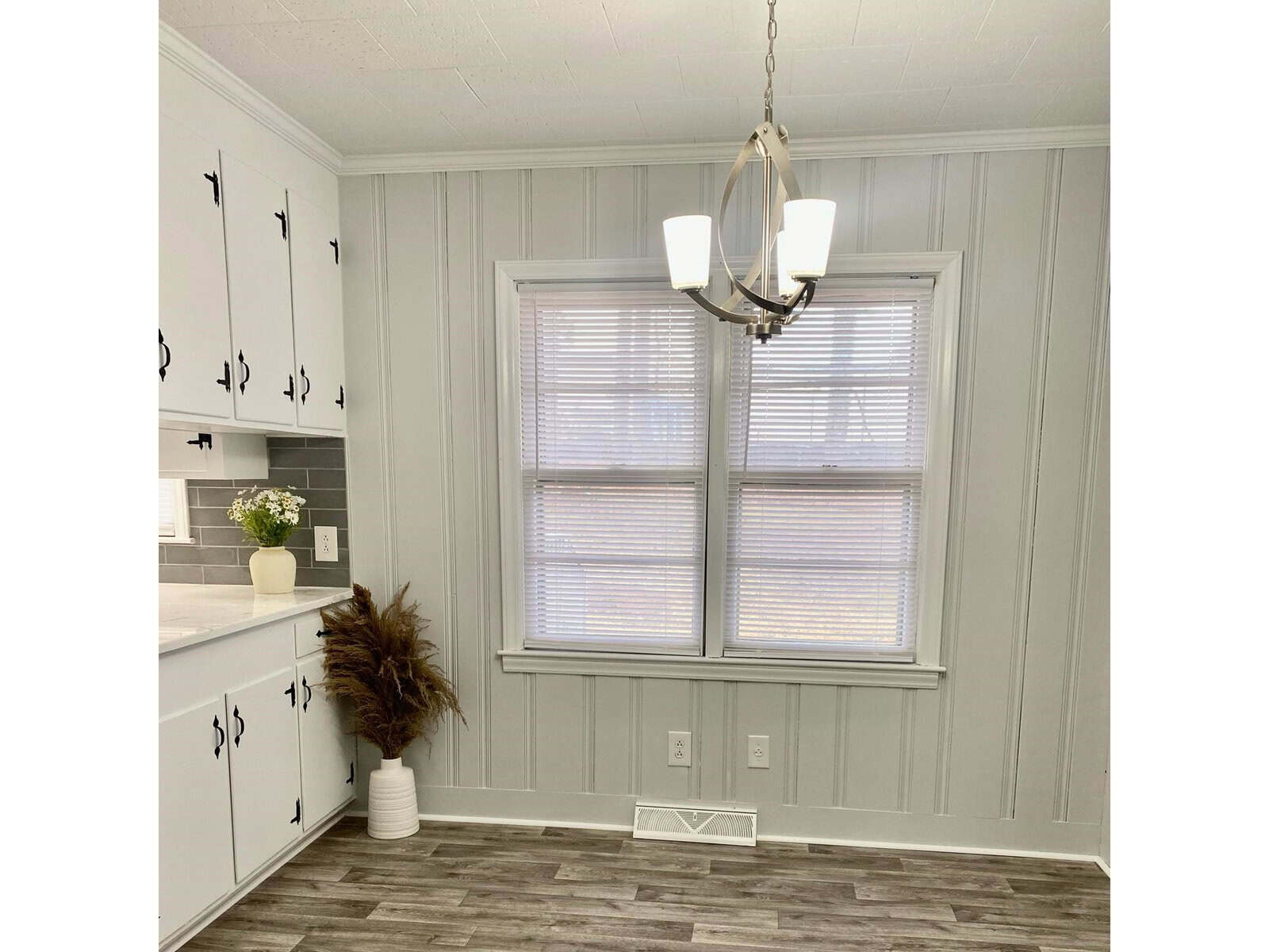 A clean and modern kitchen interior with white cabinetry, gray walls, and a chandelier hanging in front of a window with white blinds.