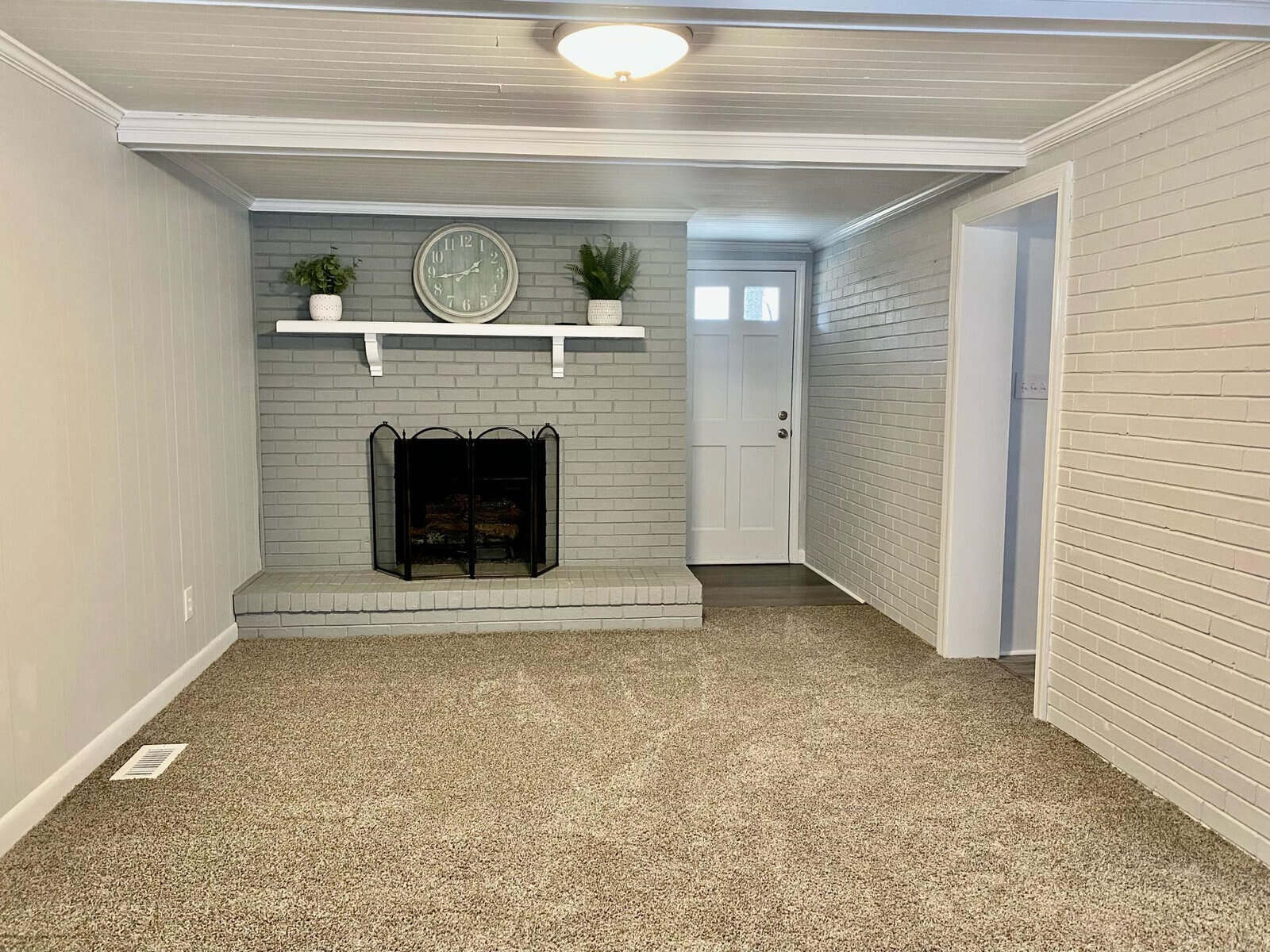 A neatly presented room with gray walls, a fireplace, a large clock above the mantel, and speckled carpet flooring. there's a white door on the right and an entrance to another room on the left.