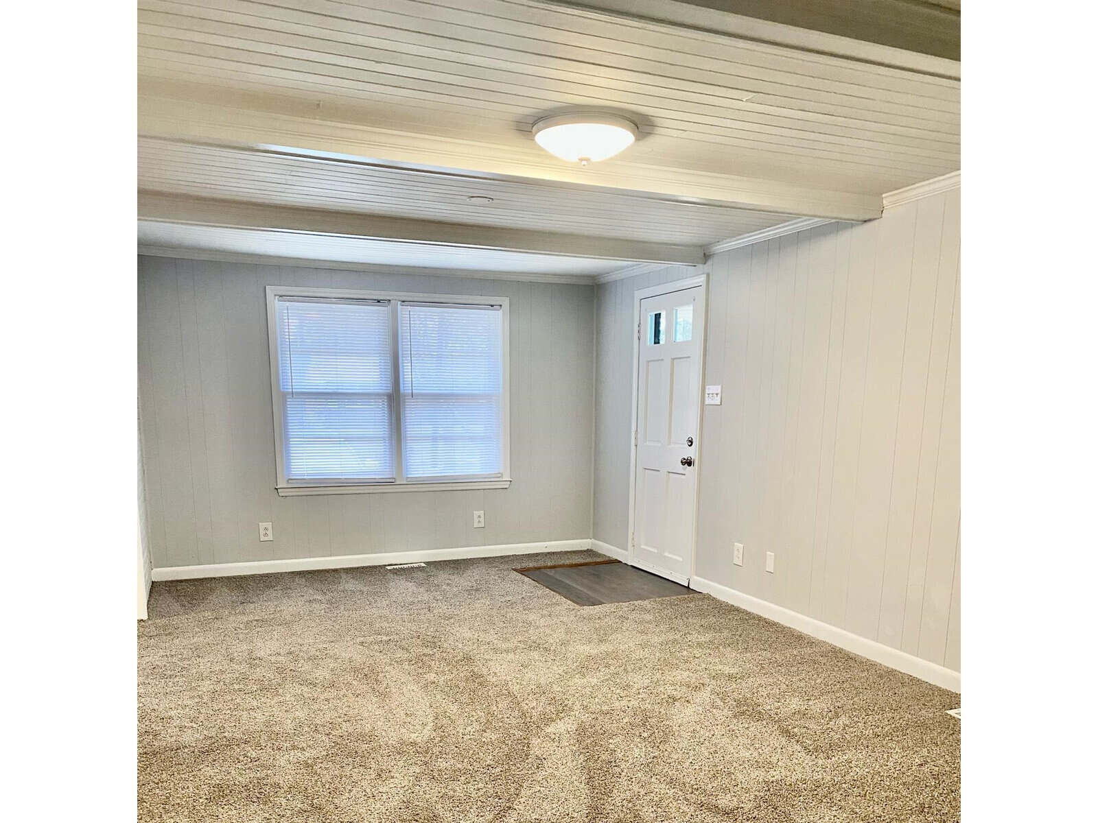 An empty room with beige carpeting, two windows with blinds, gray walls, one wall with white paneling, and a white door.