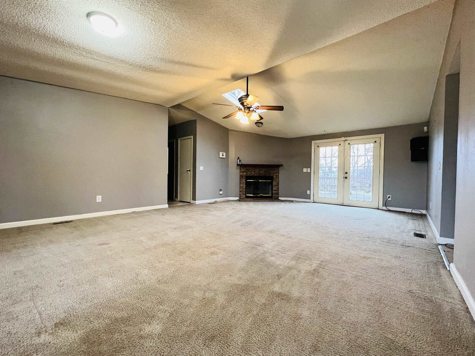 A spacious, empty living room with beige carpet and gray walls. It features a ceiling fan with lights, a brick fireplace, a skylight, and glass double doors leading outside. There's also a recessed area on one wall.
