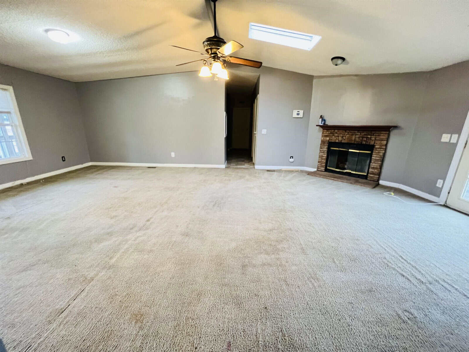 A spacious, empty living room with beige carpet and light gray walls. It features a ceiling fan with lights, a brick fireplace on the right, and a wide window on the left. The room is well-lit with natural and artificial lighting.