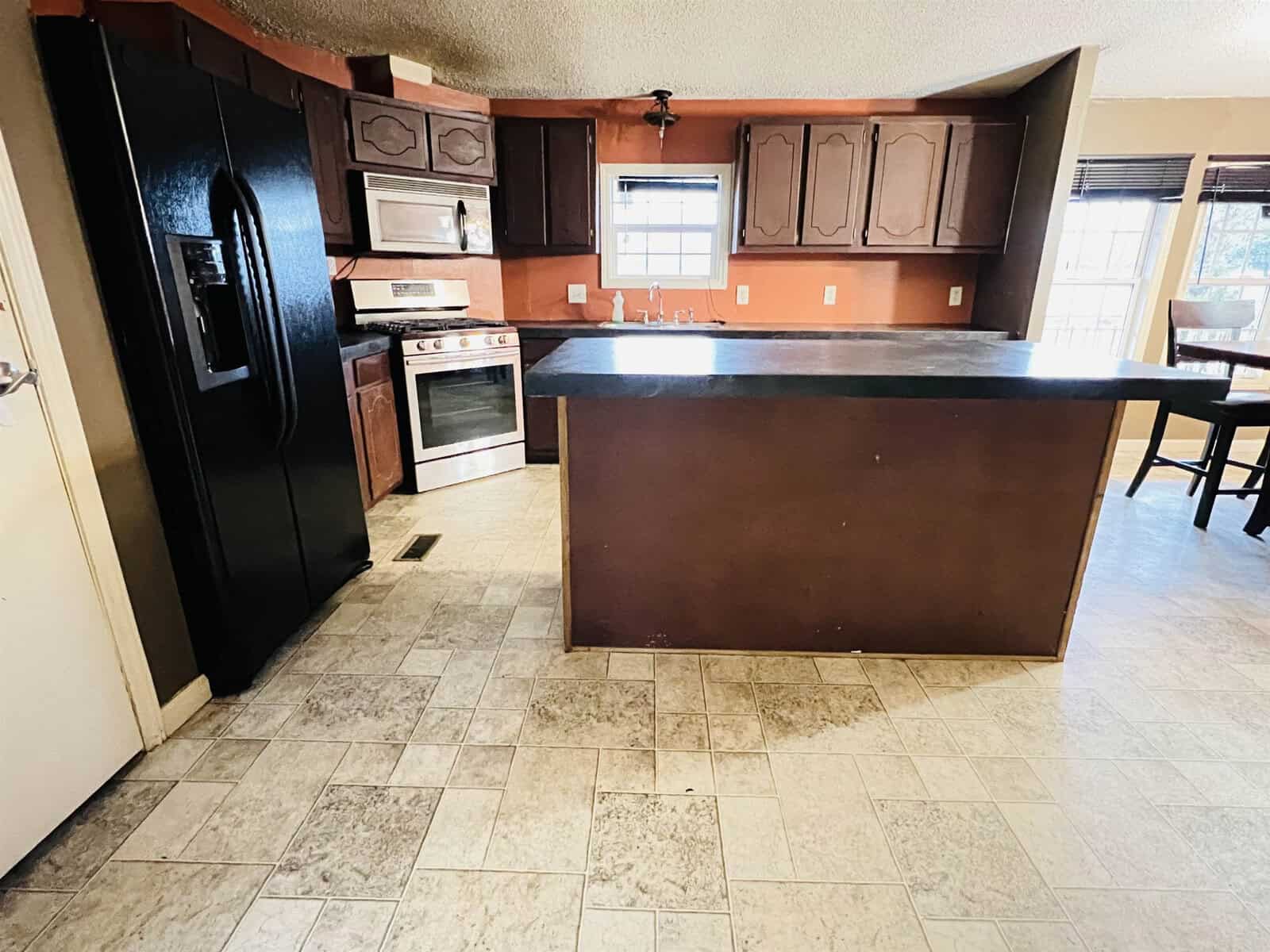 A kitchen with dark wooden cabinets, a black refrigerator, stainless steel oven, and a black countertop island. The floor is tiled, and there's natural light coming through the windows.