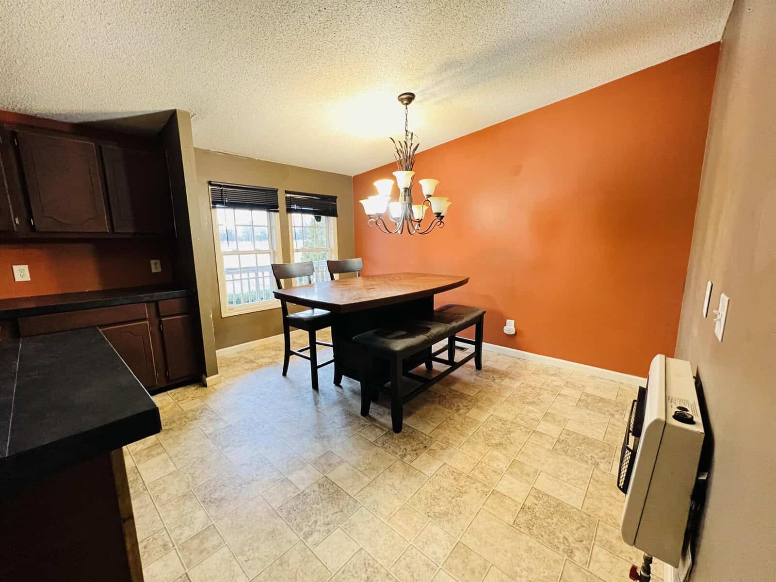 A dining room with an orange accent wall features a wooden table and two benches under a multi-bulb chandelier. Adjacent to a window with closed blinds, the room has tiled flooring and dark kitchen cabinets visible on the left.