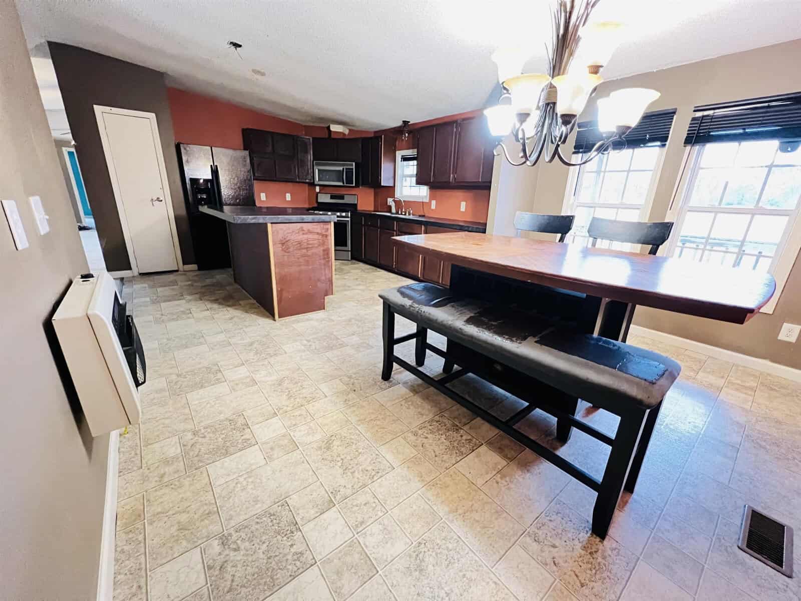 A spacious kitchen with dark wood cabinets, stainless steel appliances, and a large island with a wooden countertop. A dining table with a bench and chairs is placed on a tiled floor under a chandelier, next to large windows.