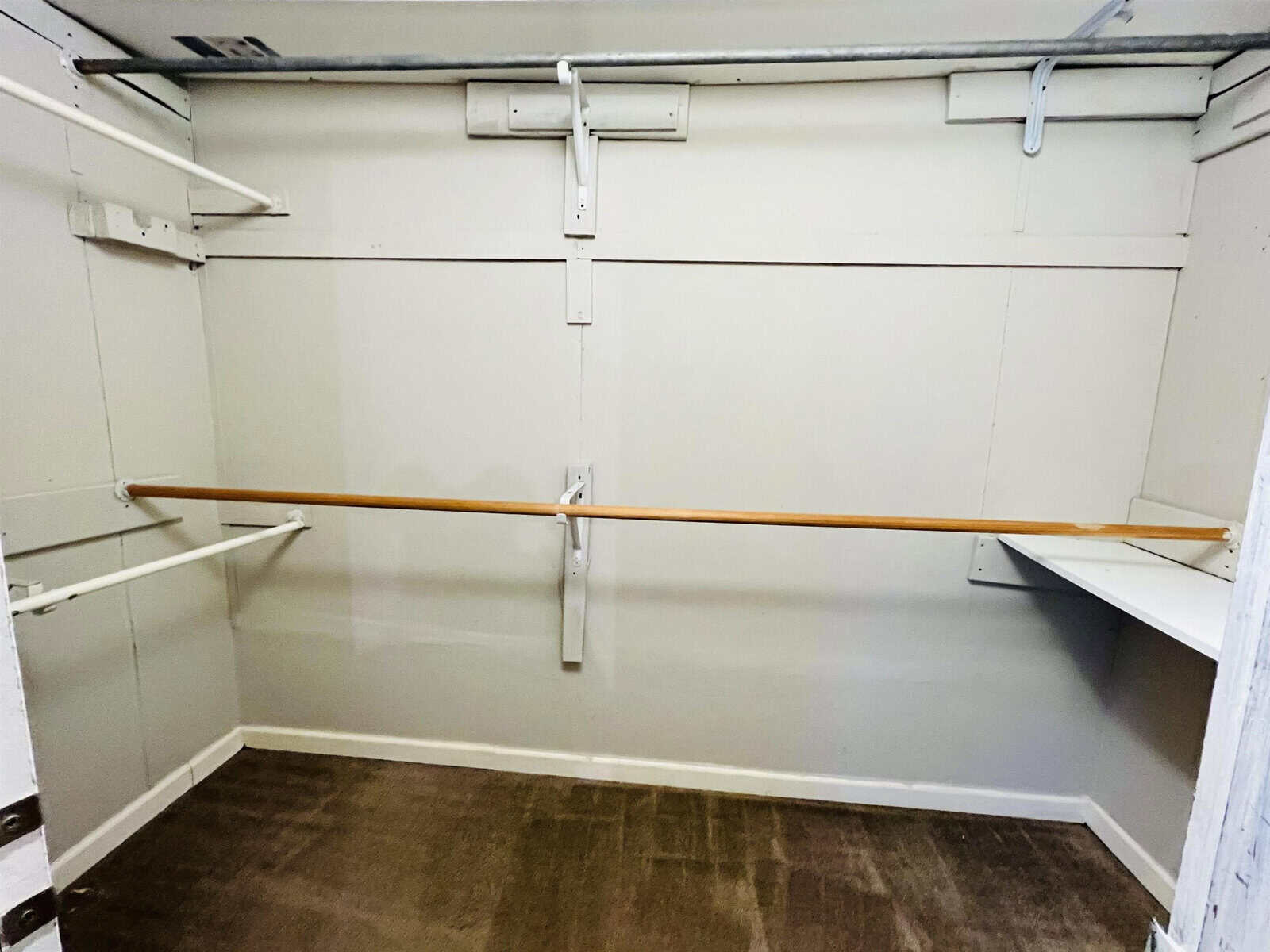 An empty walk-in closet with beige walls, a brown carpeted floor, and multiple white shelves. Wood and metal rods are installed for hanging clothes, and there is a small white built-in shelf on the right wall.