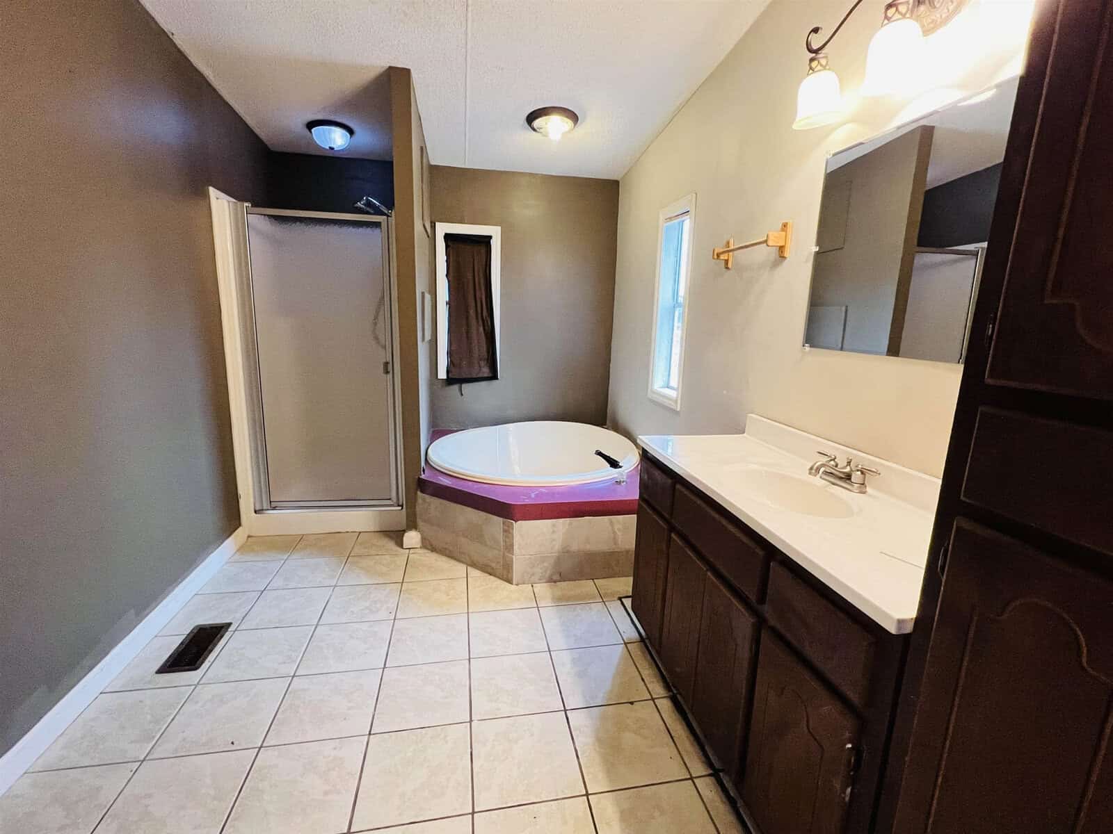 A bathroom with beige tile flooring, featuring a corner bathtub with a purple accent, a shower stall, a dark wood vanity with a white countertop, and two wall-mounted lights. Walls are painted in a dark grayish-brown color.
