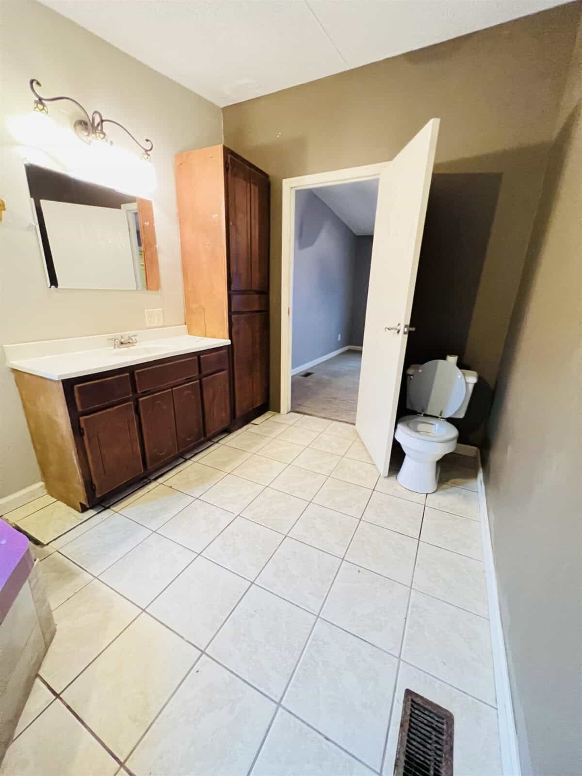 A bathroom with beige tile flooring, a wooden vanity with a sink and mirror, a toilet with the seat up, a narrow wooden cabinet, and an open door leading to an adjacent room. The walls are painted in a neutral color.