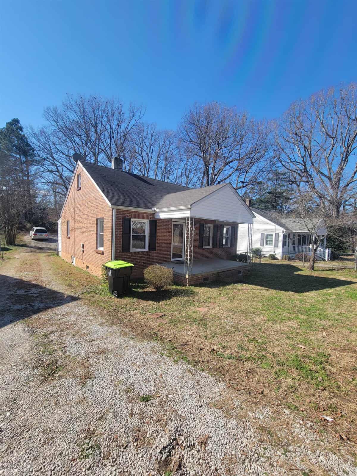 A single-story brick house with a white door and shutters, featuring a small front porch, is shown on a sunny day with a clear blue sky at 903 Dabney Drive, Henderson NC