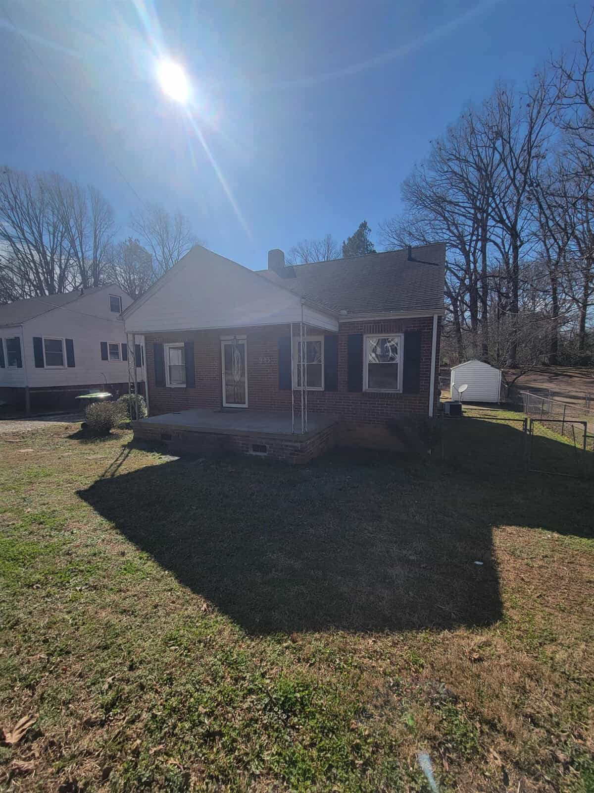 A single-story brick house with a small front porch, situated under a bright sun with a clear blue sky. the house has a gable roof and white-framed windows; there is a yard in front with no visible fencing.