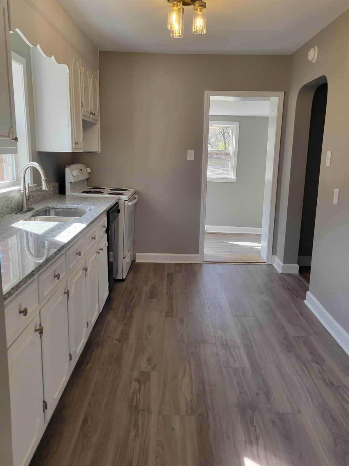 A newly renovated kitchen with white cabinetry, stainless steel appliances, and gray laminate flooring extending towards an adjoining room with a view of a window.
