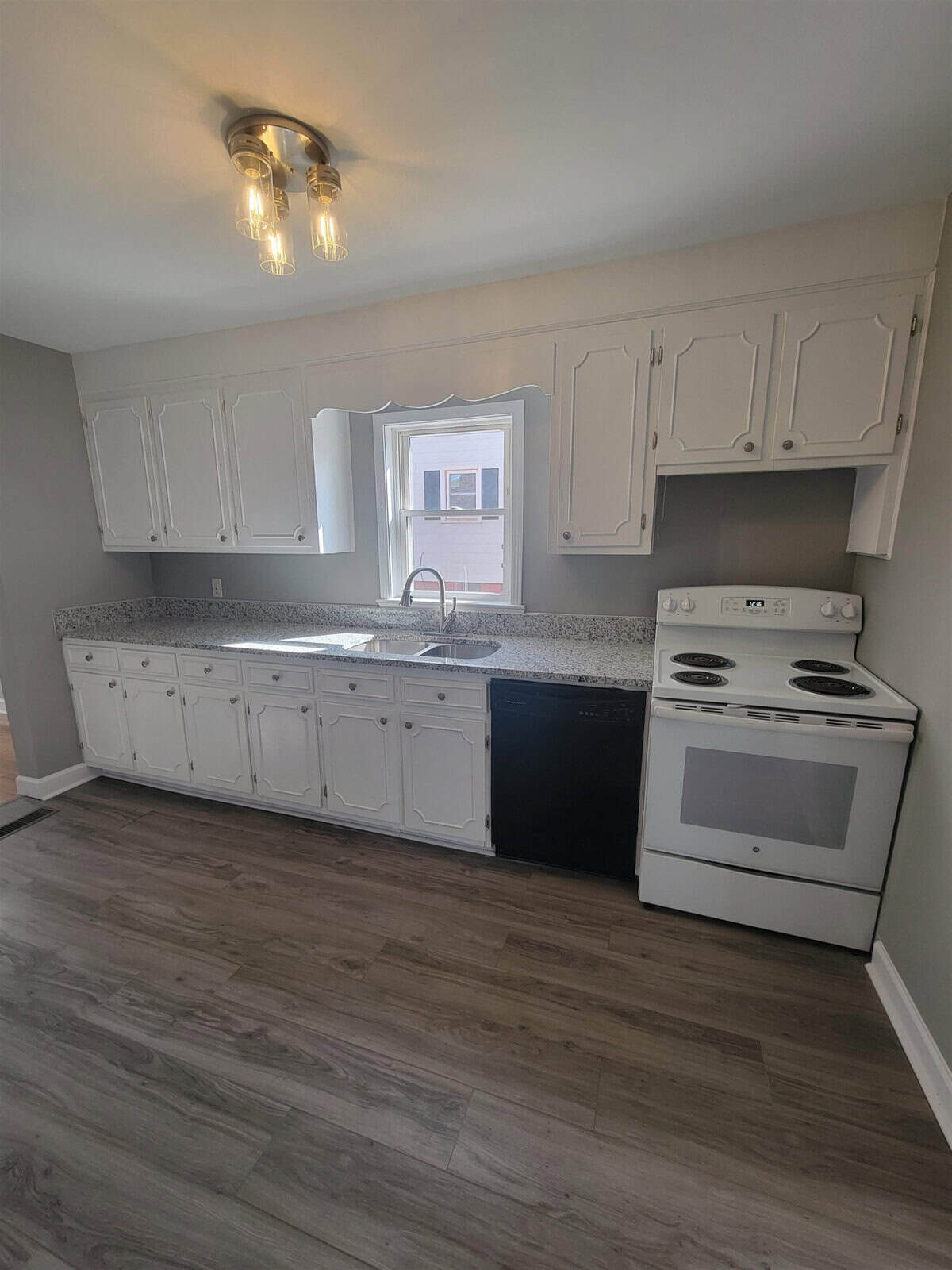 A modern kitchen interior with white cabinetry, dark countertops, and grey laminate flooring. the kitchen is equipped with a white electric stove and a dishwasher, and is well-lit by a window and overhead light fixtures.