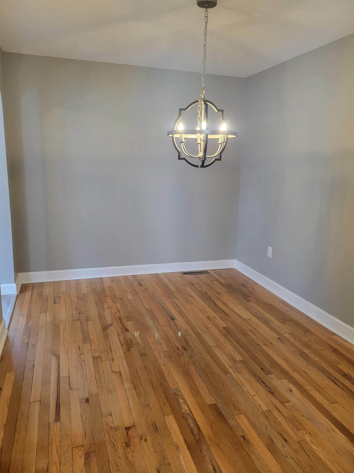 An empty room with hardwood flooring and a hanging chandelier centered in the space.