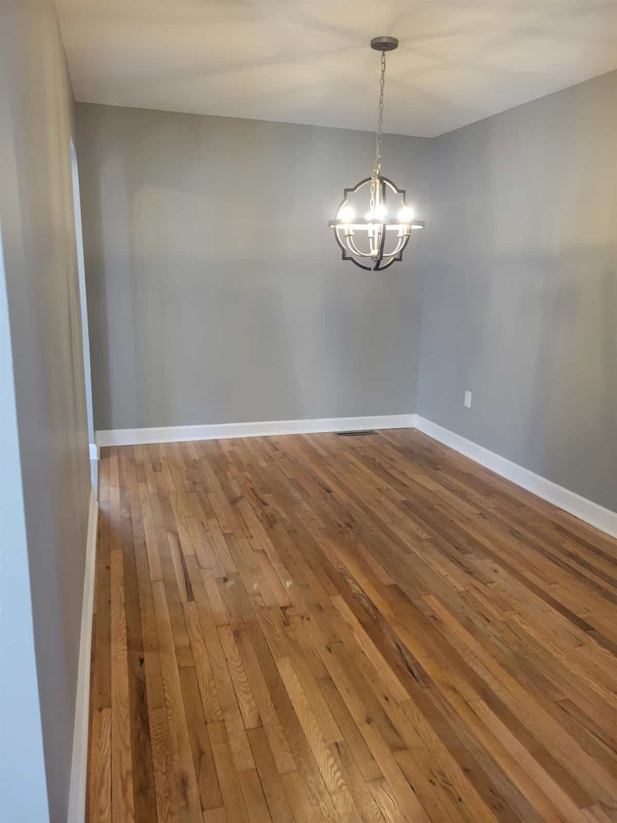 An empty room with hardwood flooring and a hanging chandelier, with walls painted in a neutral grey tone.