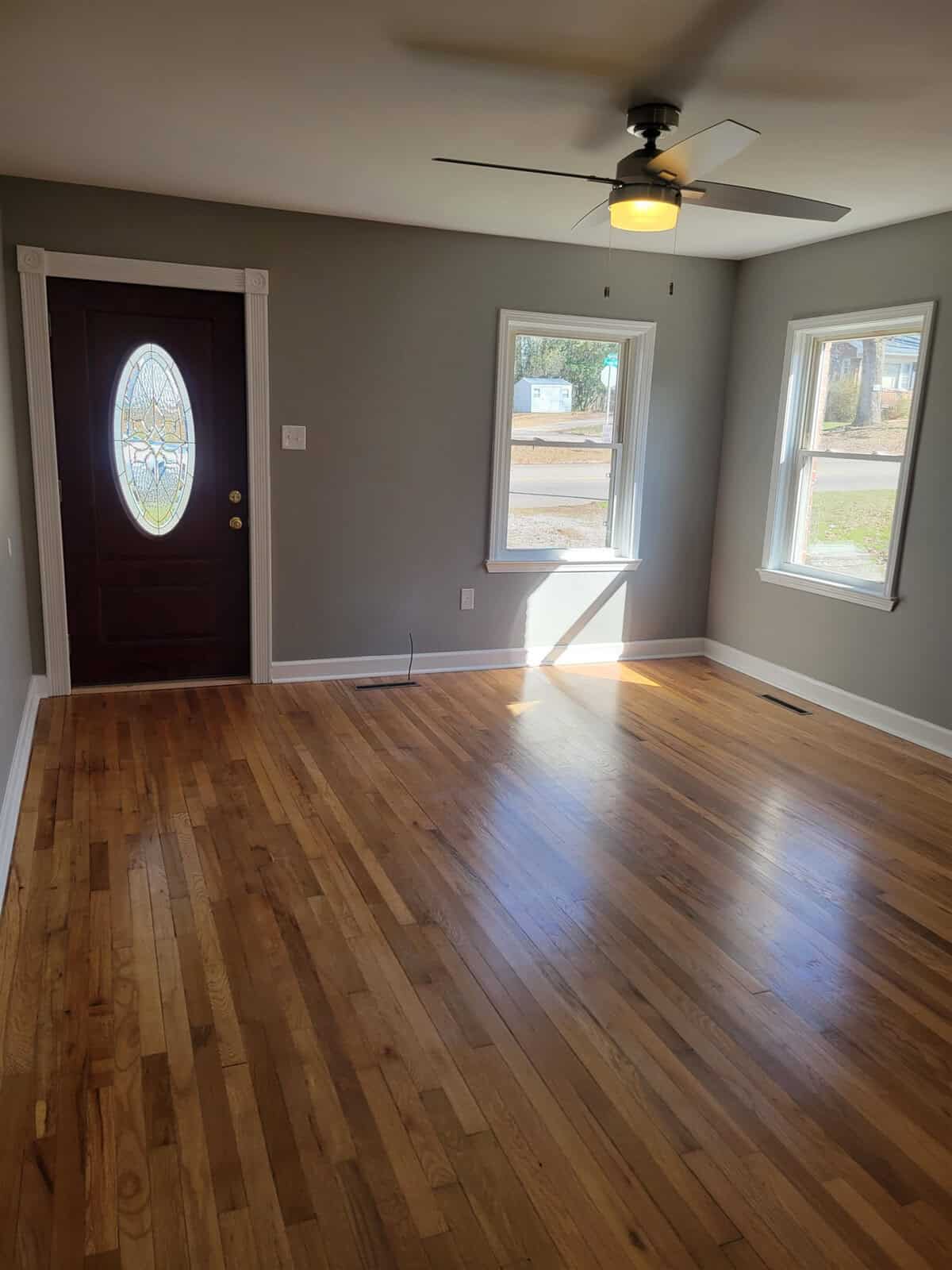 A well-lit empty room with hardwood floors, featuring a front door with an oval glass design, a ceiling fan, and two windows allowing natural light to enter.