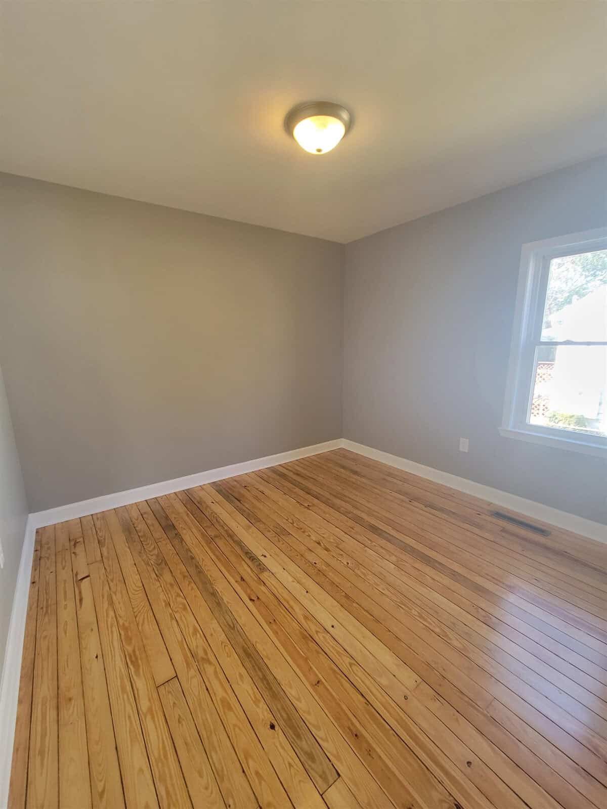 An empty room with natural wooden flooring, gray walls, a single window providing natural light, and a ceiling light fixture.
