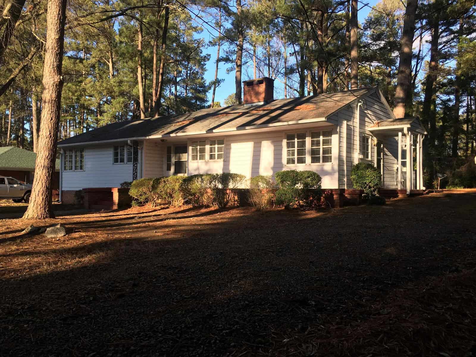 A single-story white house with shutters, surrounded by tall pine trees and covered by the shadow of the late afternoon sun.