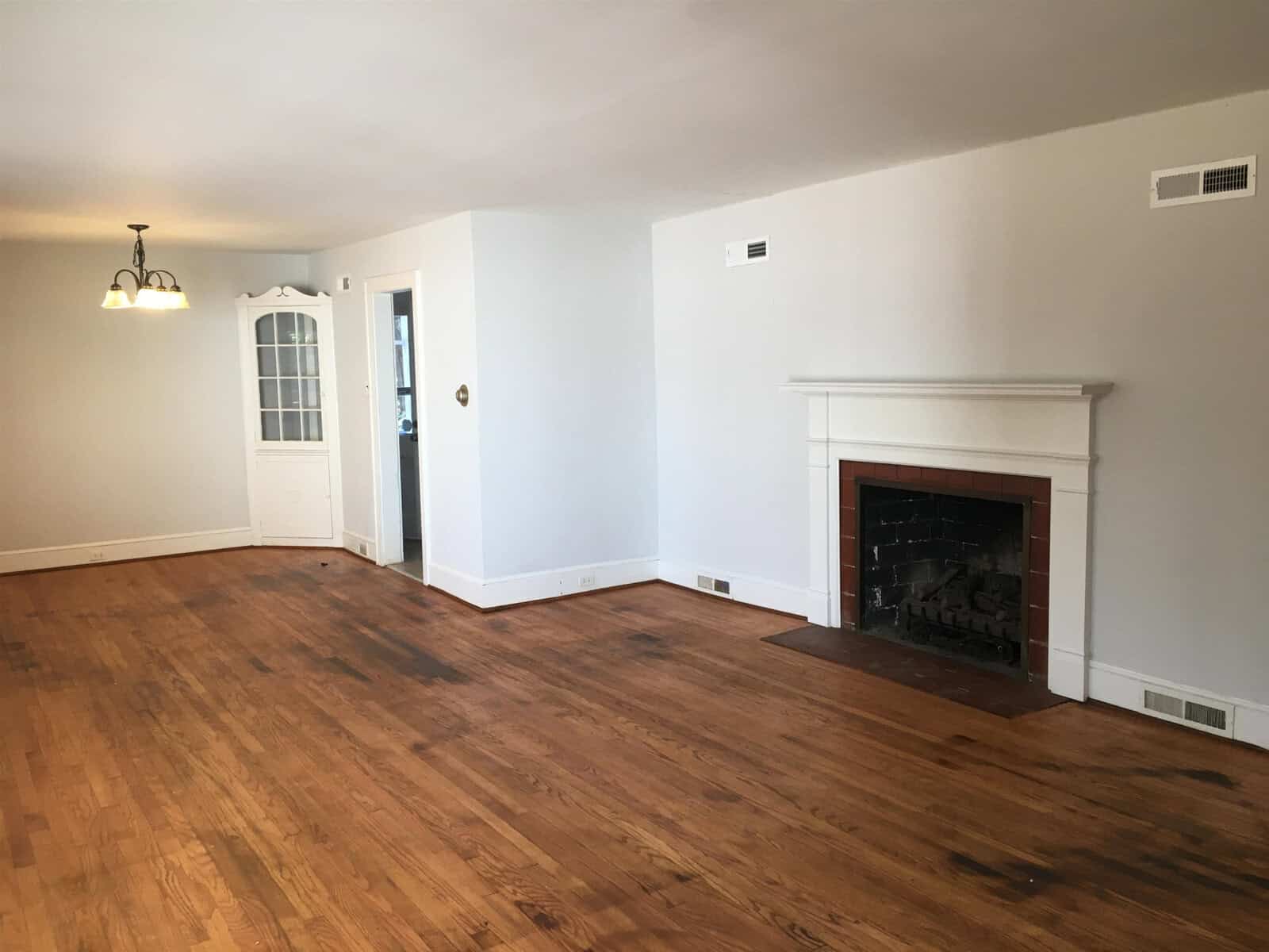 A spacious empty room featuring hardwood floors, a white fireplace on the right, and a white door leading outside on the left, with a small chandelier hanging from the ceiling.