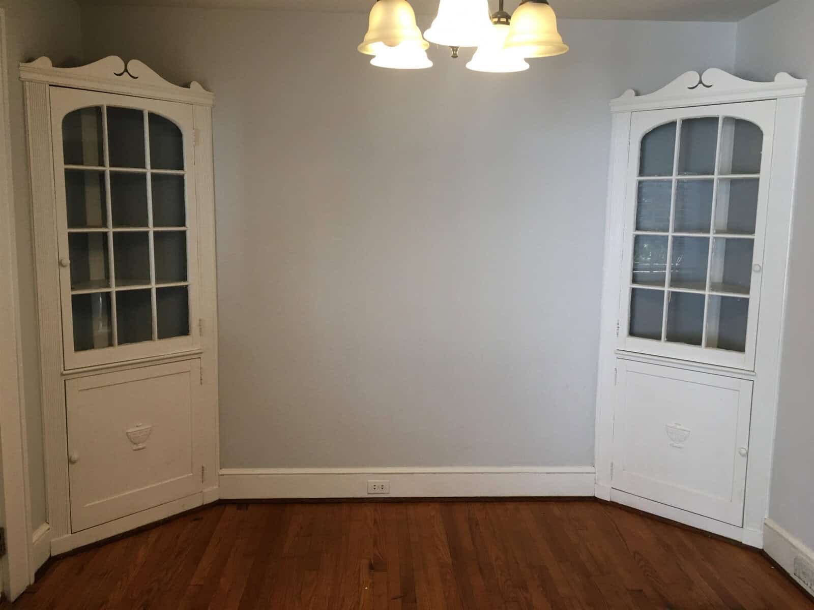 A symmetrical room featuring a wooden floor, two white corner cabinets with glass-paneled doors, and a central light fixture on the ceiling.
