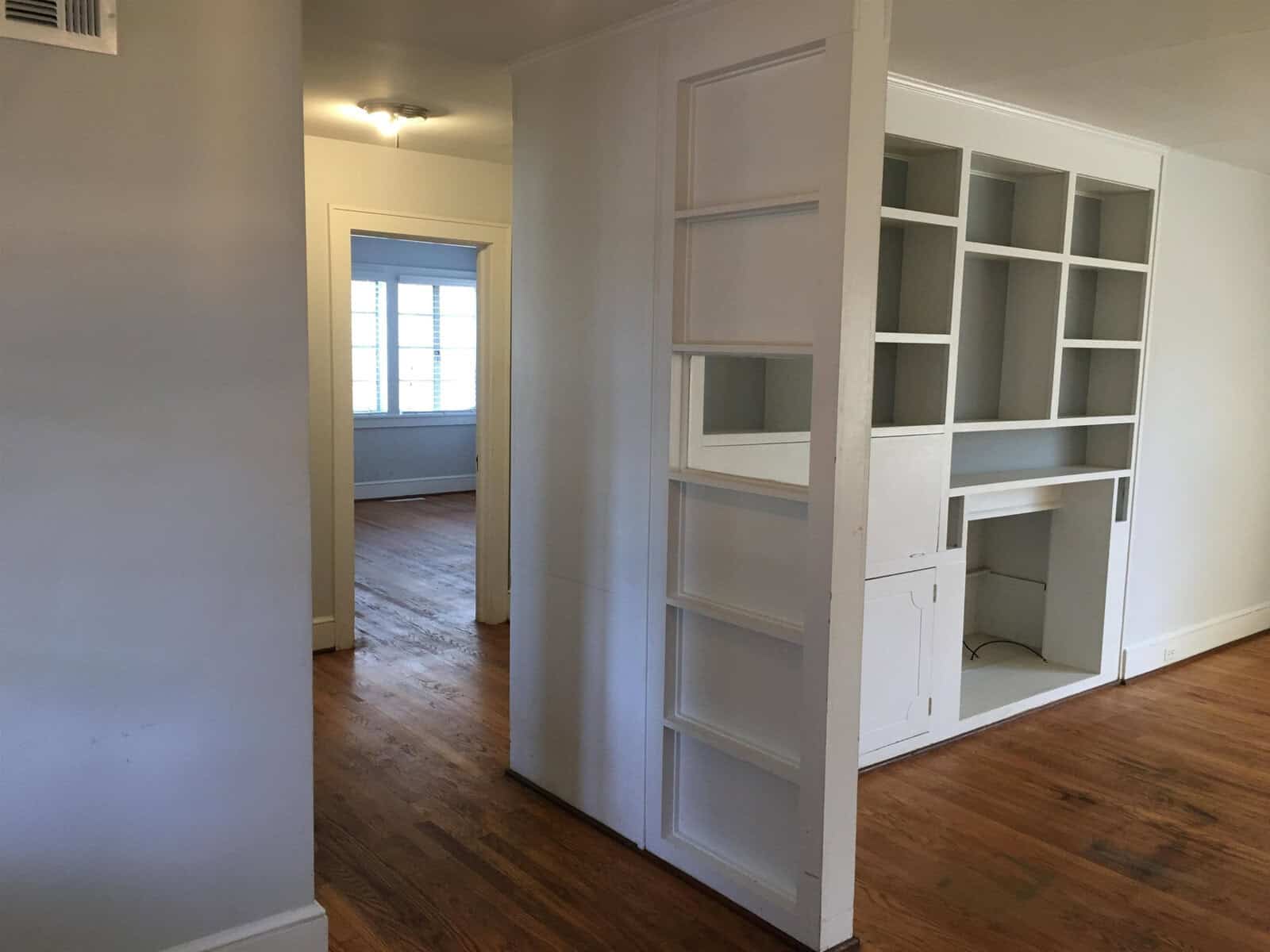 An empty room with wooden floors, an open white door, and built-in shelving units along the wall.