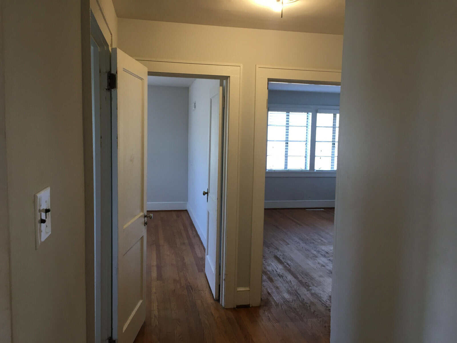 An empty room with an open doorway leading to another room, featuring hardwood floors, white walls, and closed white shutters on a window.