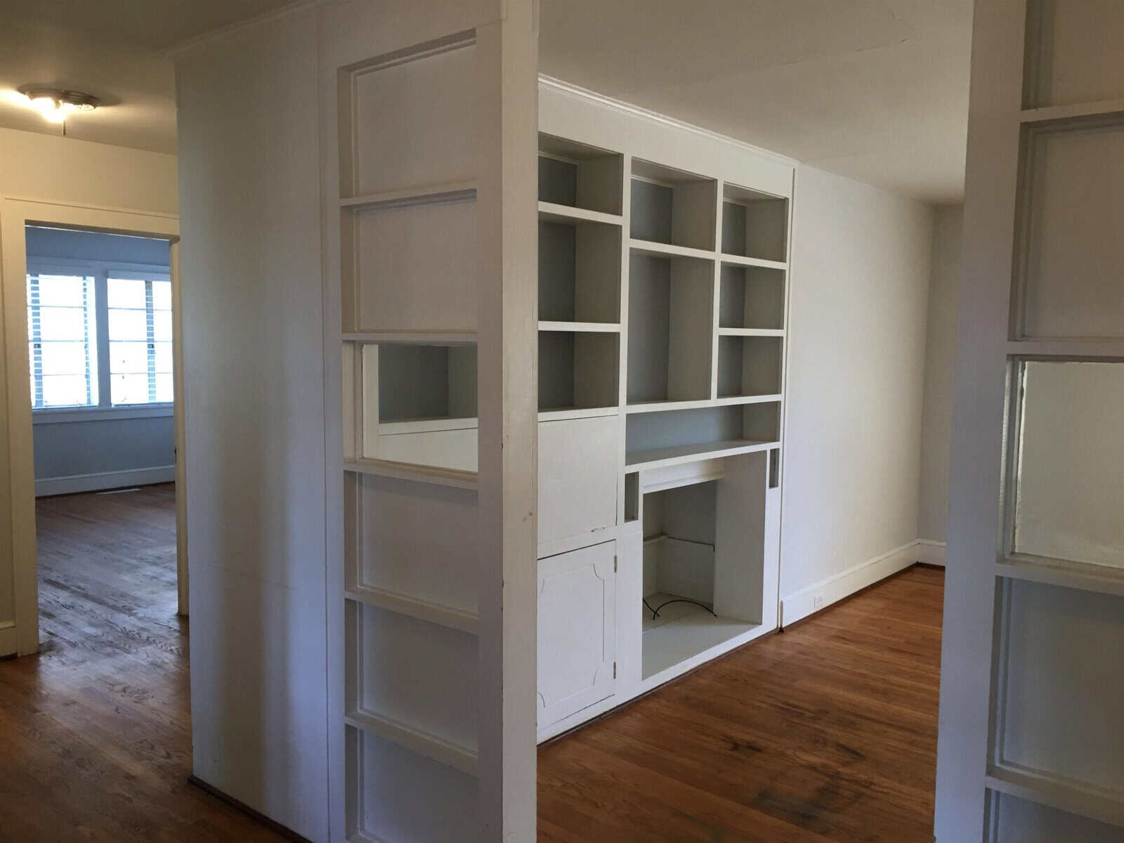 An empty room with hardwood floors featuring a built-in shelving unit and desk space, with sunlight coming through the windows.