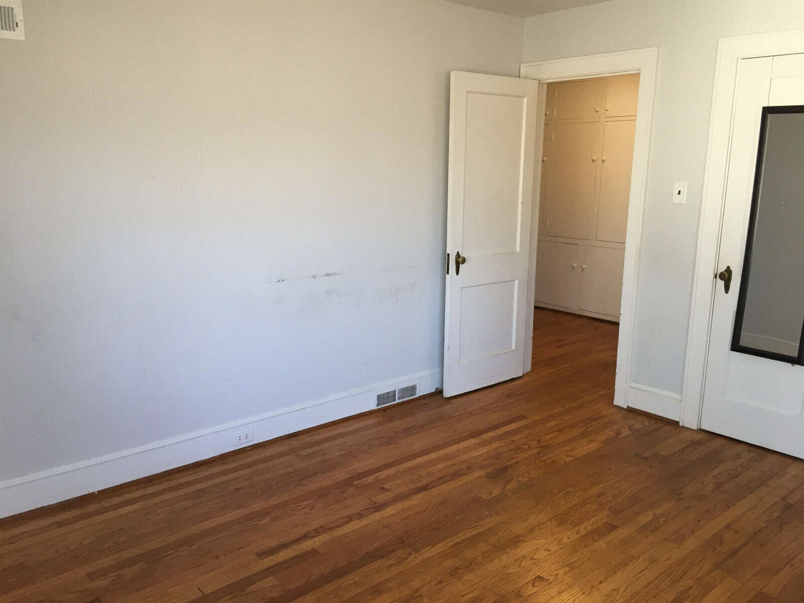 An empty room with white walls, showing signs of wear and scuff marks, with a closed white door on the left, an open closet with sliding doors in the center, and a full-length mirror on a white door to the right. the room has hardwood flooring.