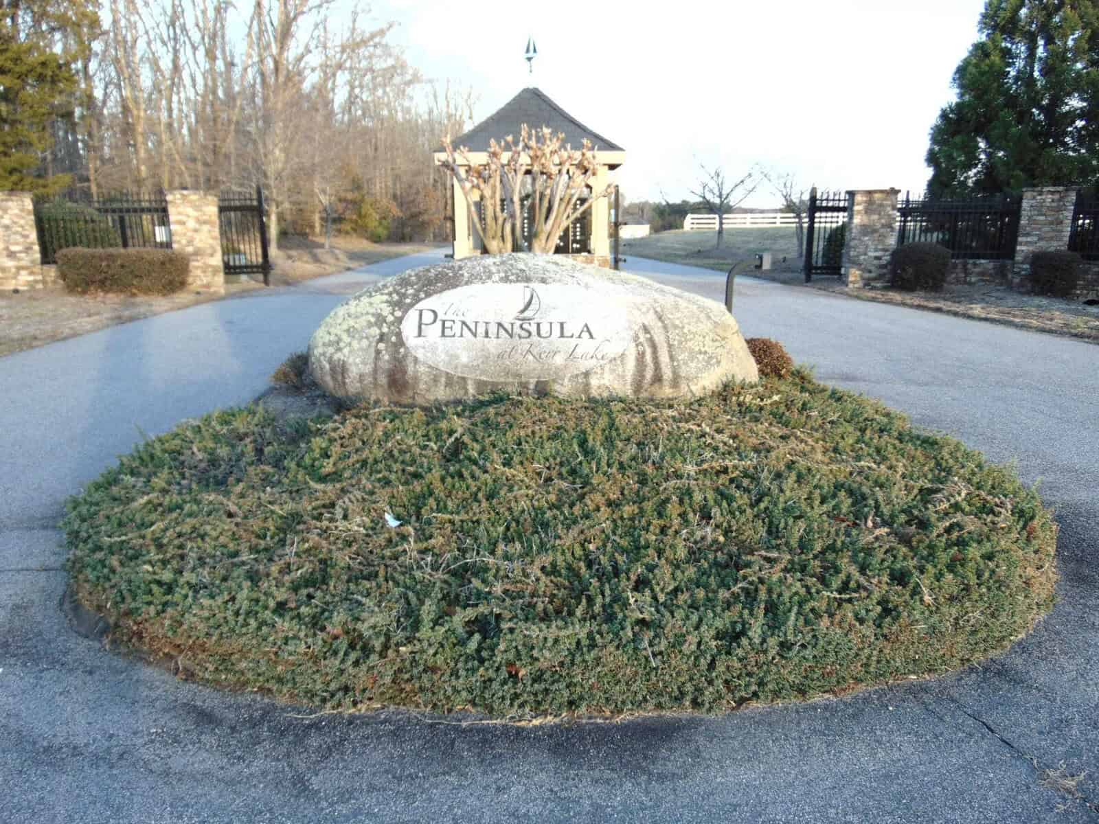 A stone sign with the inscription "peninsula" placed on a landscaped island at the entrance of a residential area during the daytime.