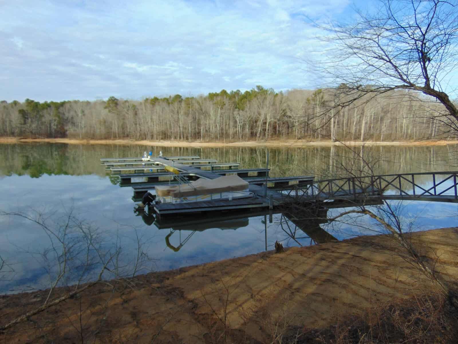 A dock with a boat moored on a tranquil lake, surrounded by bare trees under a clear blue sky.