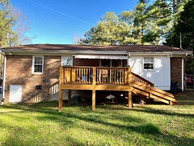 A single-story brick and white siding house with a wooden deck and stairs in the front yard, surrounded by trees with a clear blue sky above.