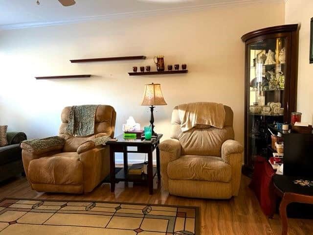 A well-lit living room featuring two beige recliners with a side table and lamp between them, a decorative rug on the floor, and a cabinet with collectibles to the right. floating shelves with small decorative items are mounted on the wall above.