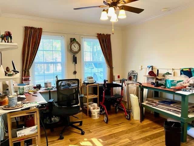 A home office and craft room with organized clutter, featuring a desk, rolling chairs, shelves with supplies, and a walker for mobility assistance.
