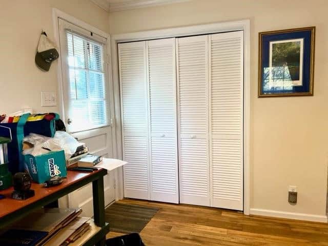 A room corner with a white closet, a cluttered desk containing various items, a window with blinds partially opened, and a framed picture on the wall.