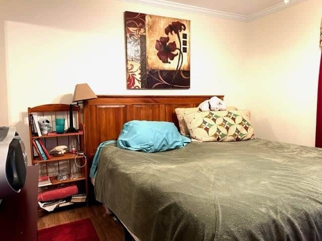 A neatly made bed with a green blanket and blue pillow in a room with a wooden headboard, a floral painting above, and a shelf containing various items and books to the left.