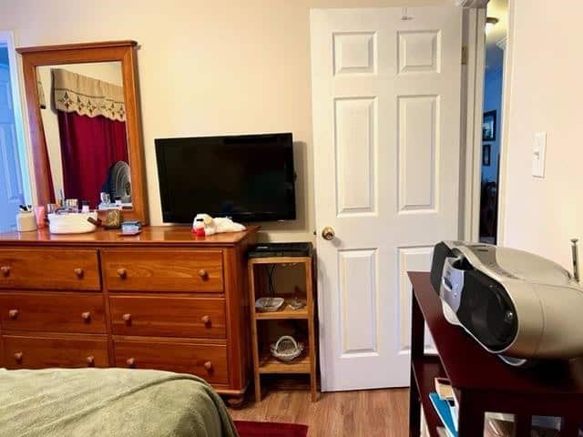 A bedroom interior featuring a wooden dresser with a mirror, a flat screen tv, a white door, and a corner of a bed with a green cover. a portable radio sits on a bedside table to the right.