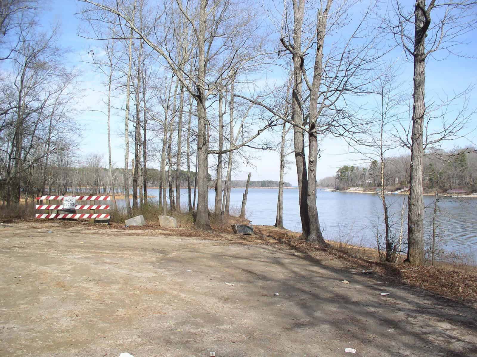 A barricade blocks a pathway leading to a tranquil lake surrounded by bare trees, with a clear sky above.